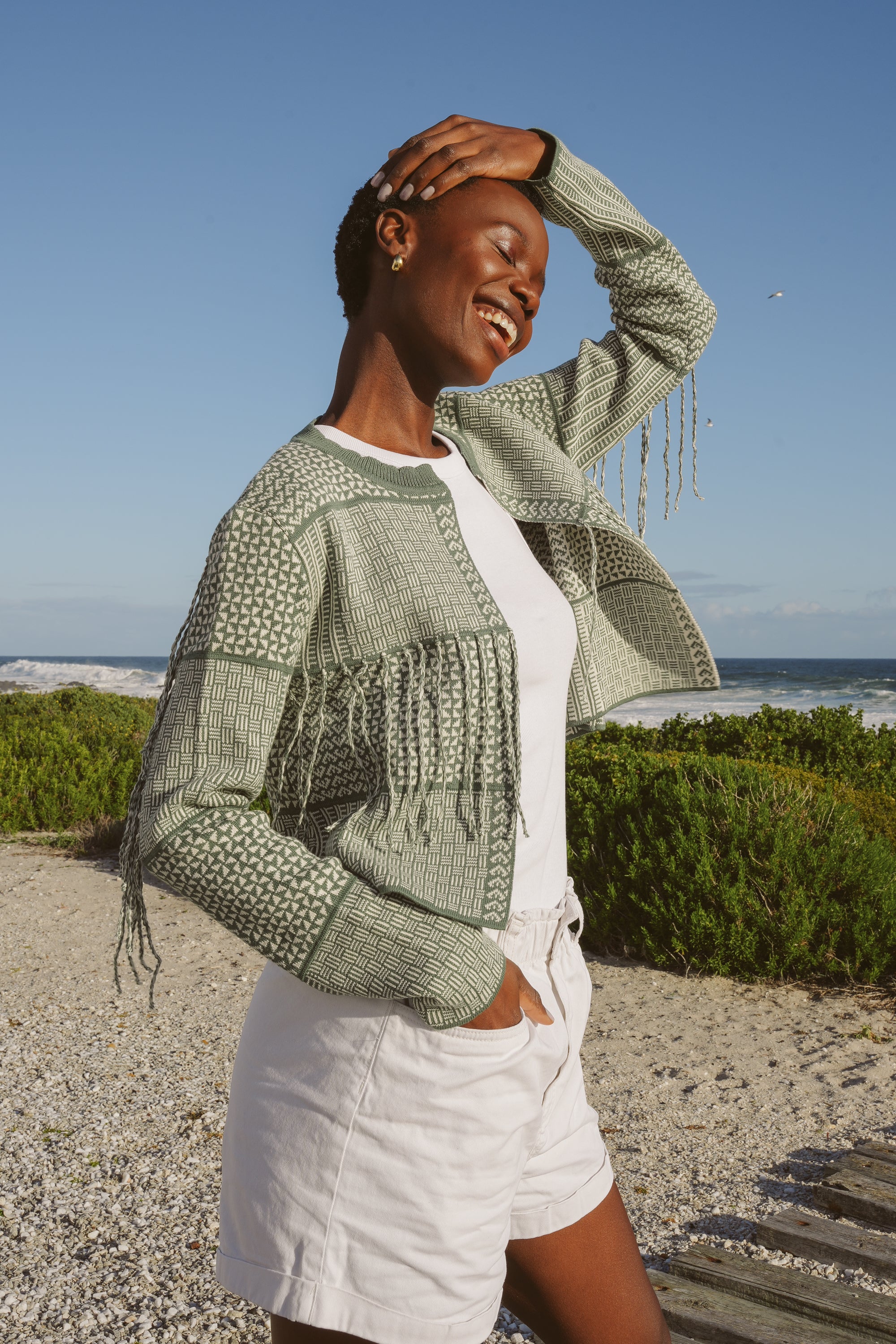 Model, Zana, wearing The Zuri knitted cardigan in Sage & Ecru, white tank top, and white shorts, on a beach.