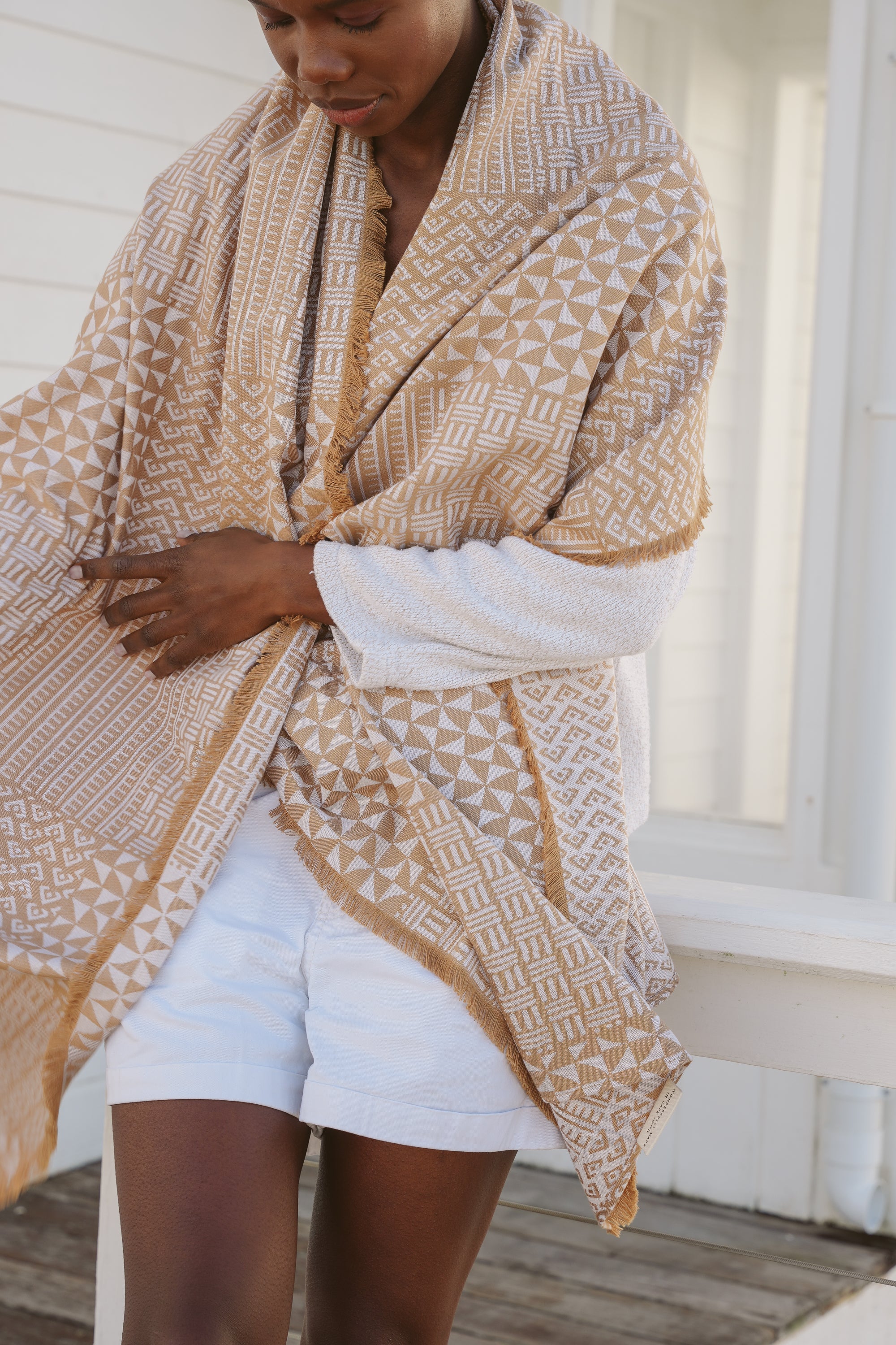Model, Zana, wearing a Zuri patterned Camel and Ecru Njeri Shawl over a ecru sweater and shorts, standing on a wooden deck.