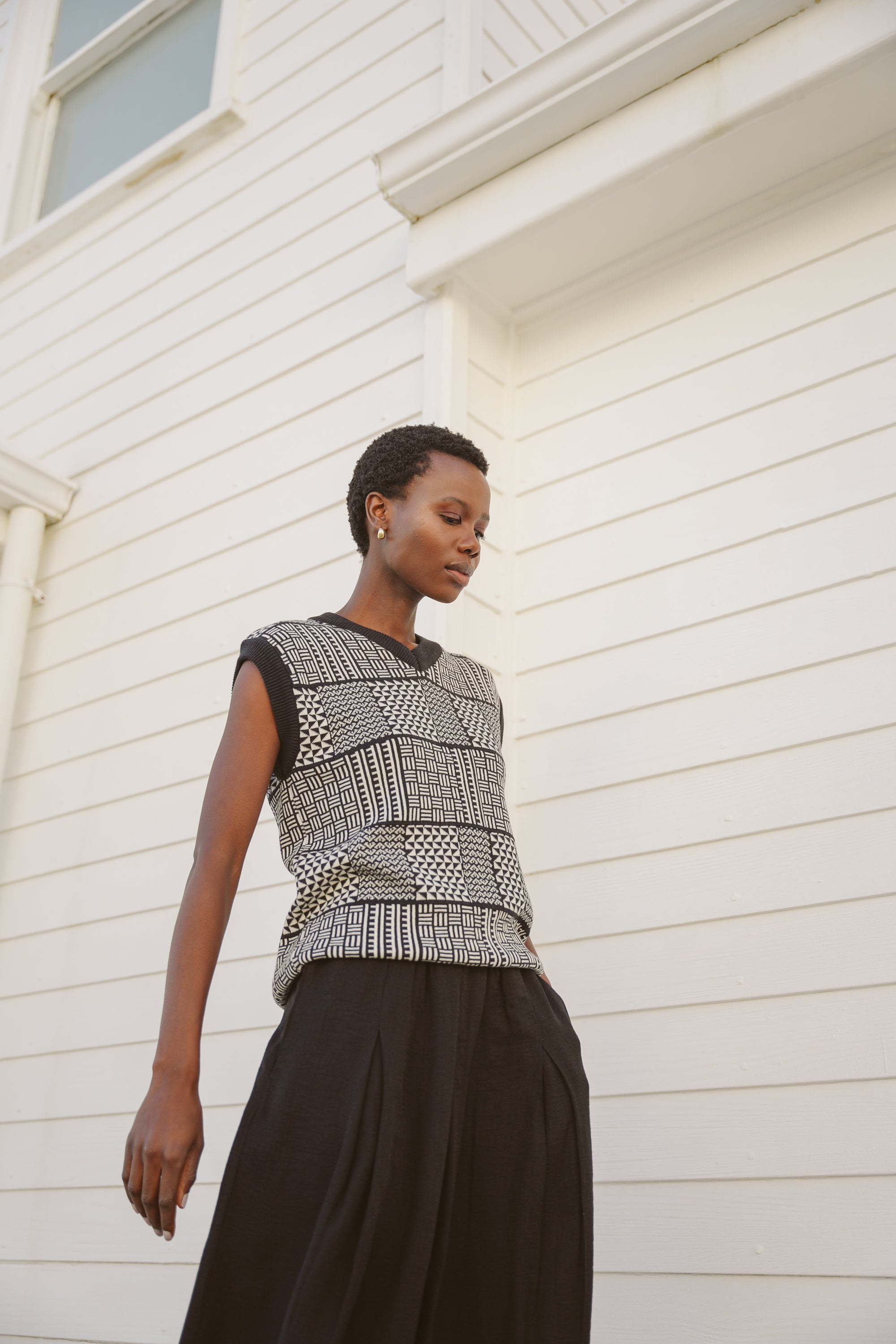 Model, Zana, wearing the sleeveless black & ecru Ade Vest with Zuri pattern, and black box pleated skirt standing against the facade of a white beach house.