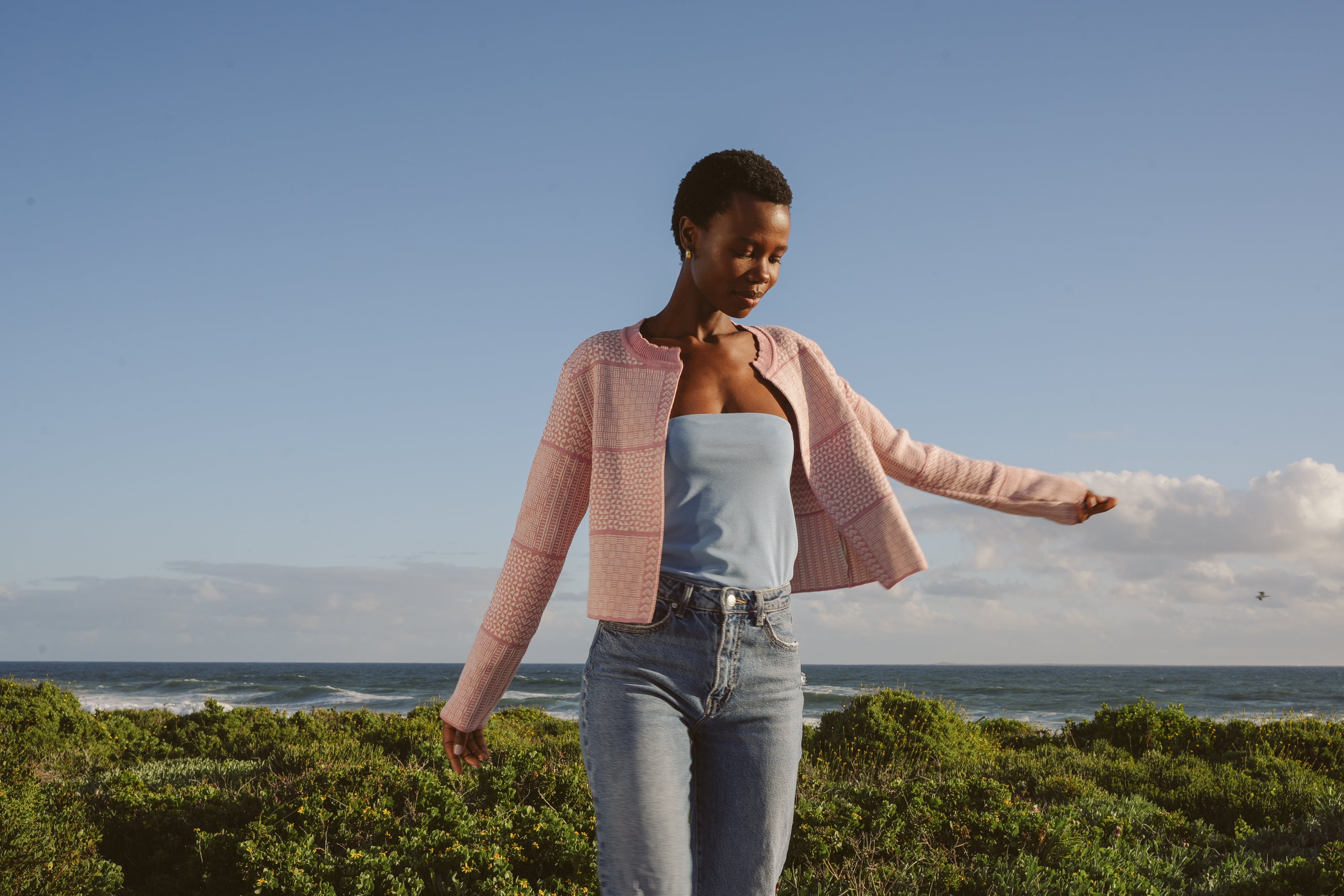 Model, Zana, wearing Dusty Pink & Ecru Fringless Zuri Cardigan. Standing on a grassy area with ocean and sky in the background.