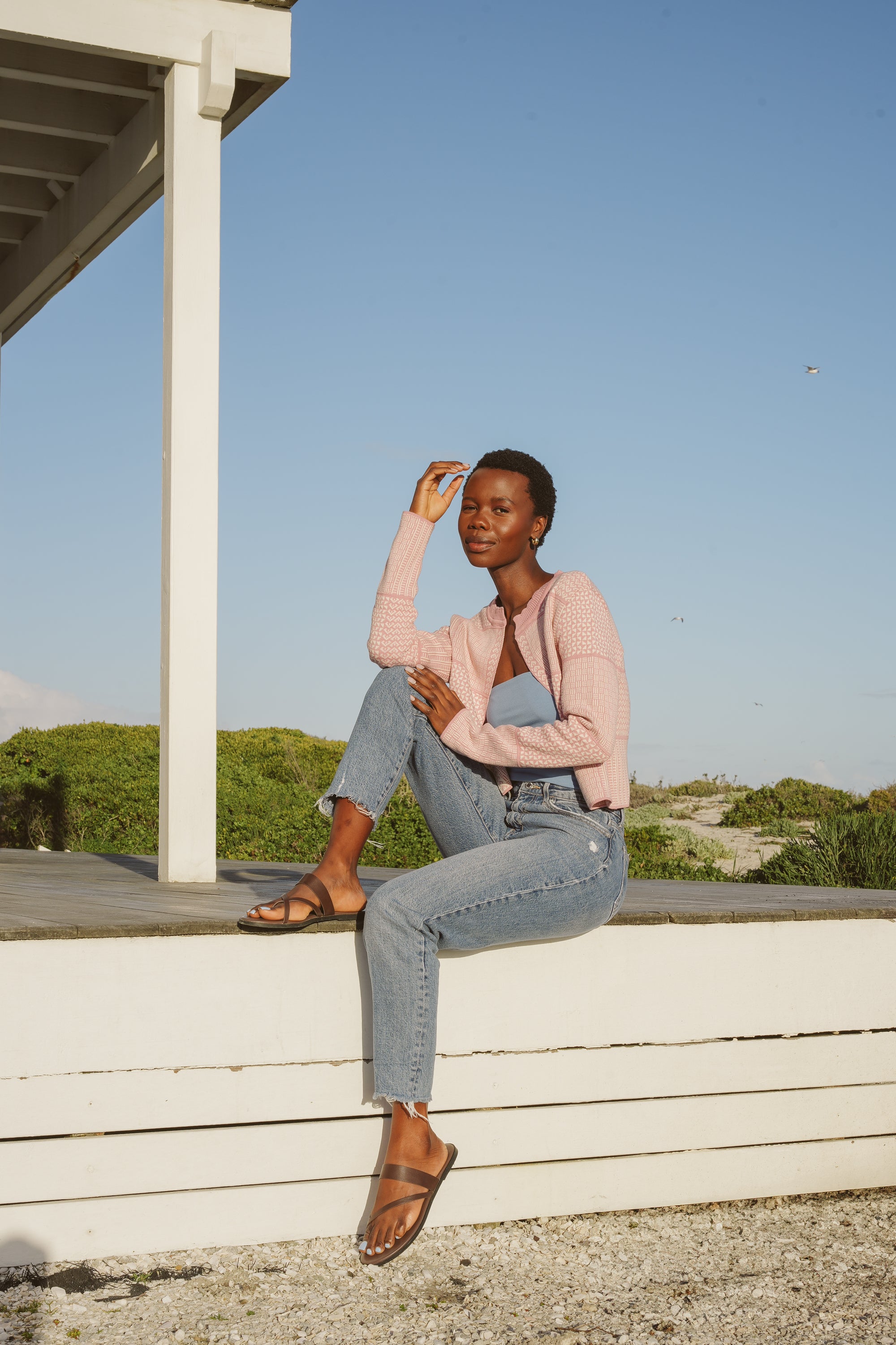 Model, Zana, wearing Dusty Pink & Ecru Fringless Zuri Cardigan. light blue top, jeans, and sandals, sitting on a ledge outdoors with a clear blue sky.